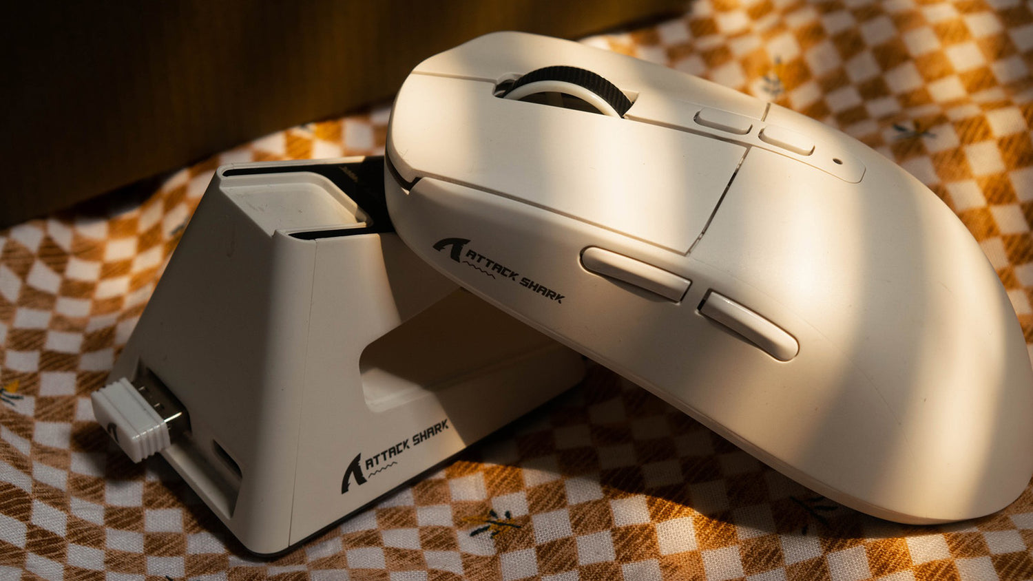 A white Attack Shark gaming mouse sits beside its charging dock on a patterned surface